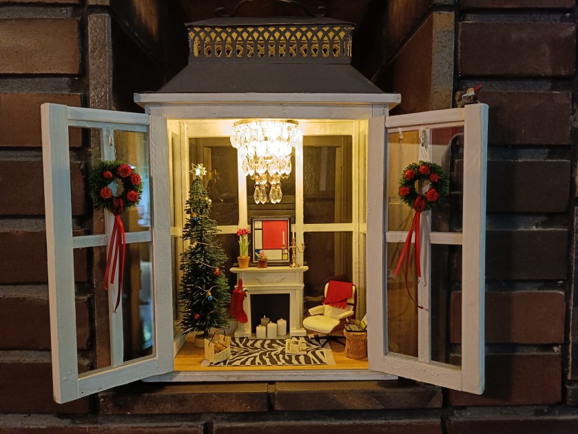 Iso kynttilälyhty, johon on rakennettu jouluinen pienoismaailma olohuoneesta.