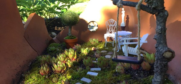 Isoon rikkinäiseen ruukkuun tehty keijupuutarha, jossa on keinu, ja pari tuolia. Alustana kasvaa sammalta ja pieniä mehikasveja.