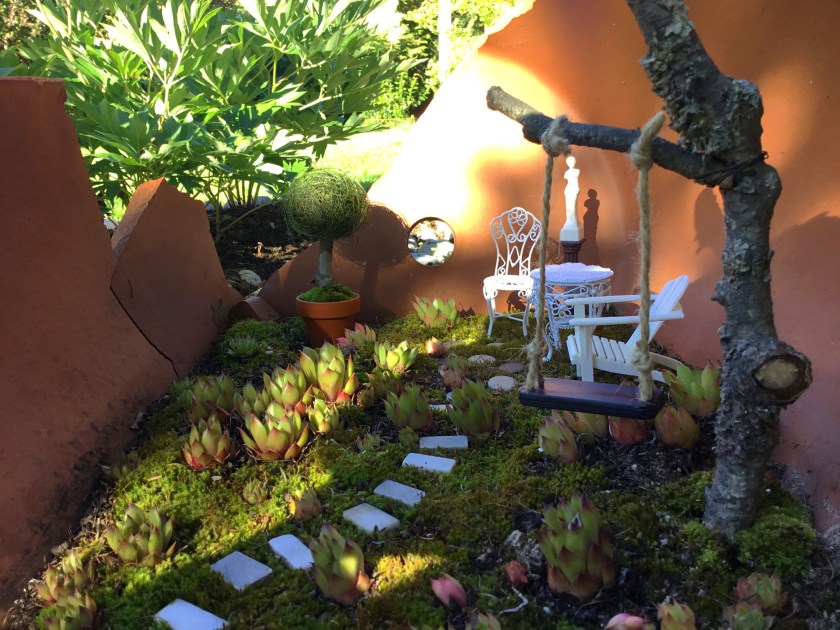 Isoon rikkinäiseen ruukkuun tehty keijupuutarha, jossa on keinu, ja pari tuolia. Alustana kasvaa sammalta ja pieniä mehikasveja.