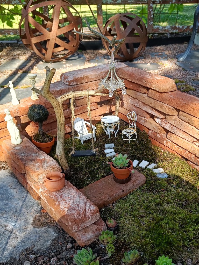 Laattatiilistä keijuille koottu puutarha, jossa keinu, istuinryhmä, veistoksia ja ruukkuja.