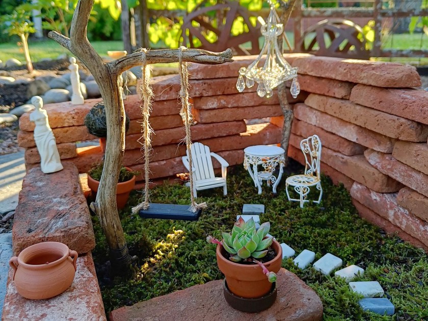 Laattatiilistä keijuille koottu puutarha, jossa keinu, istuinryhmä, veistoksia ja ruukkuja.