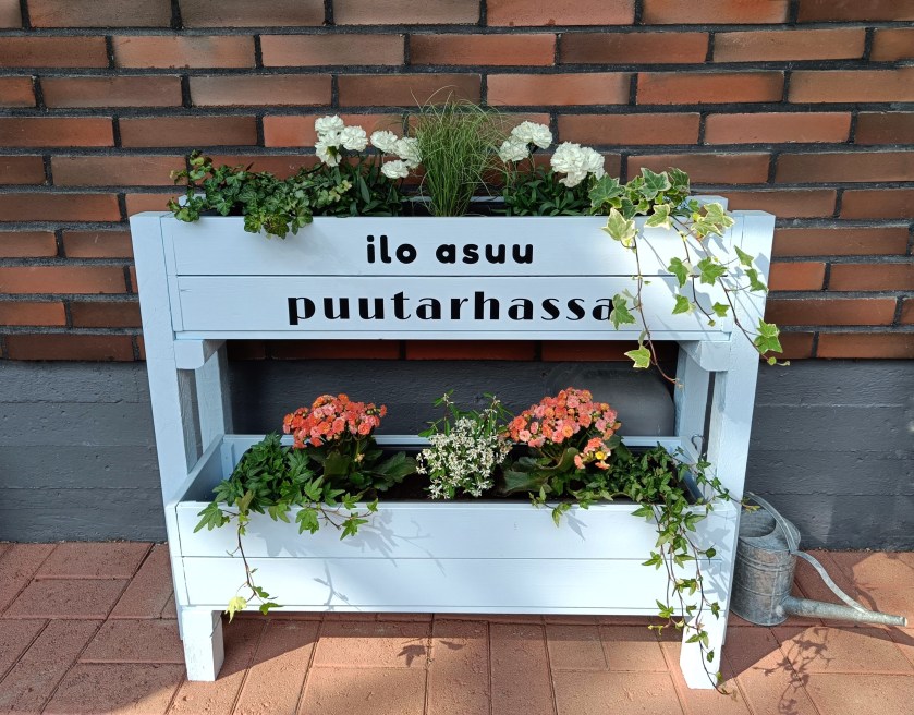Puisista parvekelaatikoista koottu kaksikerroksinen kukkateline, jossa teksti ”ilo asuu puutarhassa”.